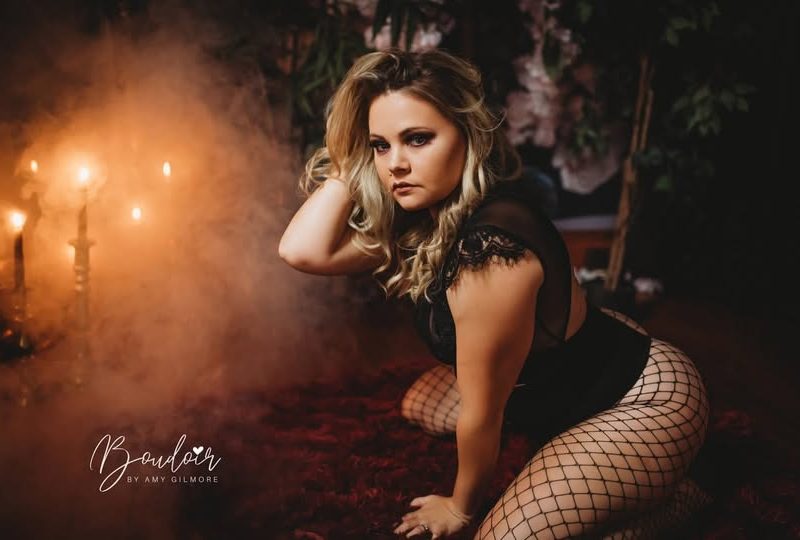 Woman in fishnet stockings kneels on a dark red rug, surrounded by a moody setting with soft candlelight and smoke, conveying elegance and intimacy.