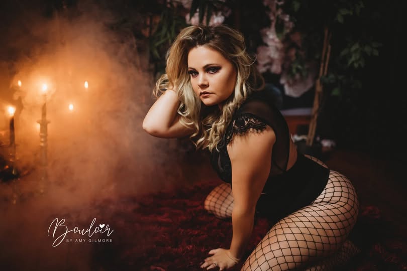 Woman in fishnet stockings kneels on a dark red rug, surrounded by a moody setting with soft candlelight and smoke, conveying elegance and intimacy.