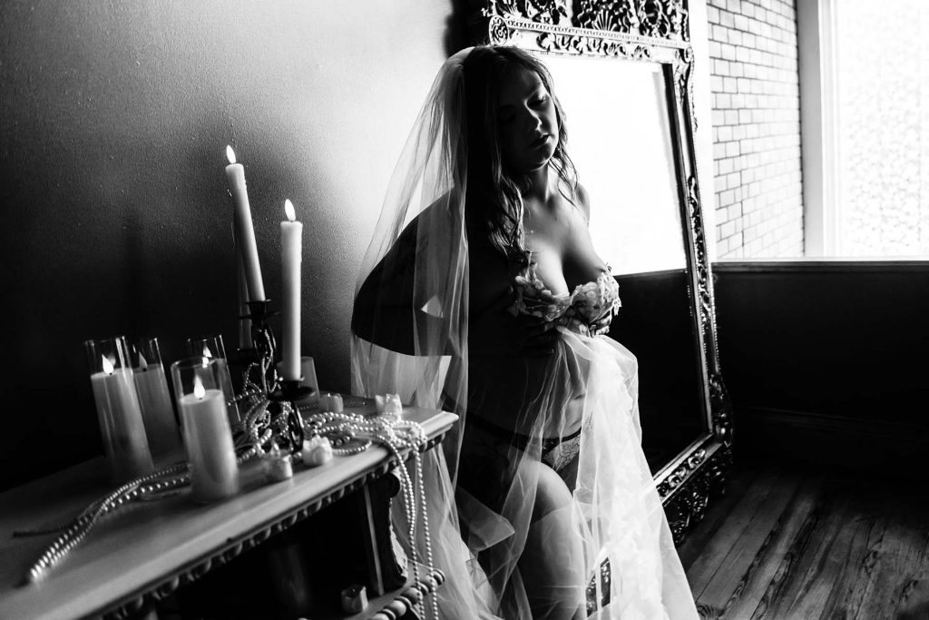 A woman in lingerie and veil poses sensually by a large ornate mirror. Nearby, a table holds tall candles and pearl necklaces. The mood is intimate.
