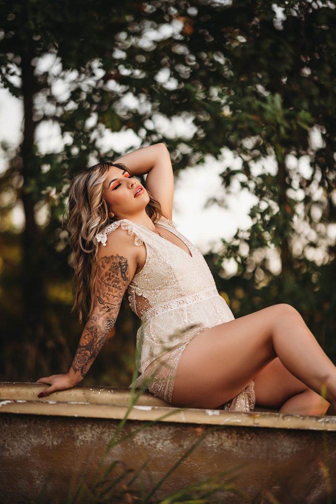 A woman in a lace romper leans back on an old bathtub outdoors, with a forested background. Her arm is decorated with tattoos, conveying a serene and confident mood.