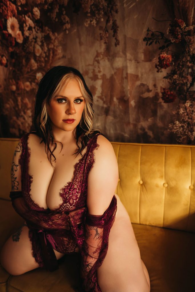 Woman in burgundy lace lingerie sits on a yellow sofa, set against a floral backdrop, exuding confidence and elegance.