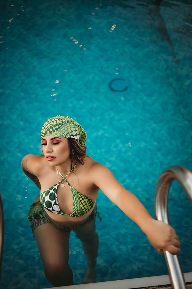 A woman in a green patterned bikini and headscarf climbs pool stairs, eyes closed, exuding calm and relaxation against the vibrant blue water.