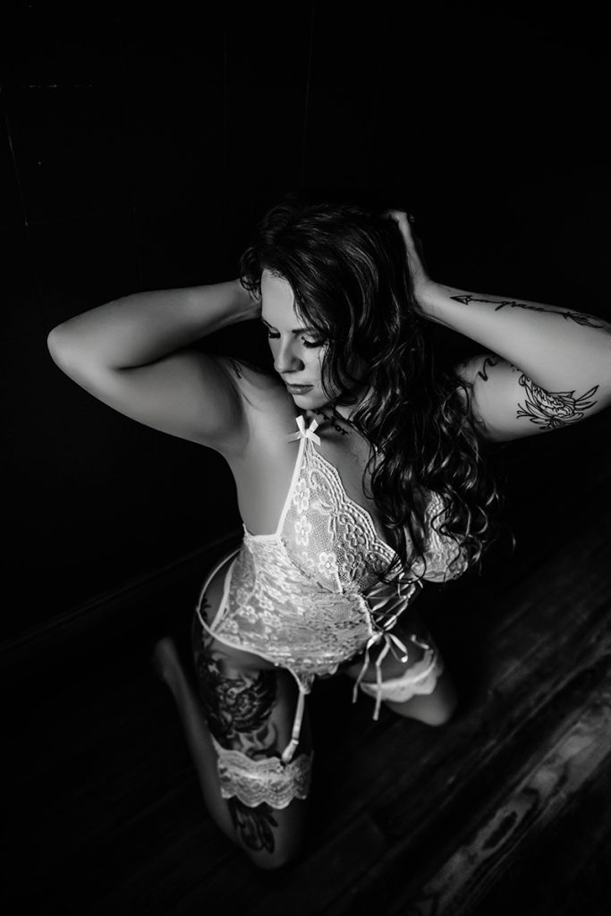 A woman kneels on a wooden floor in a dimly lit room, wearing lace lingerie. Her arms are raised, and she exudes confidence and elegance. Black and white.