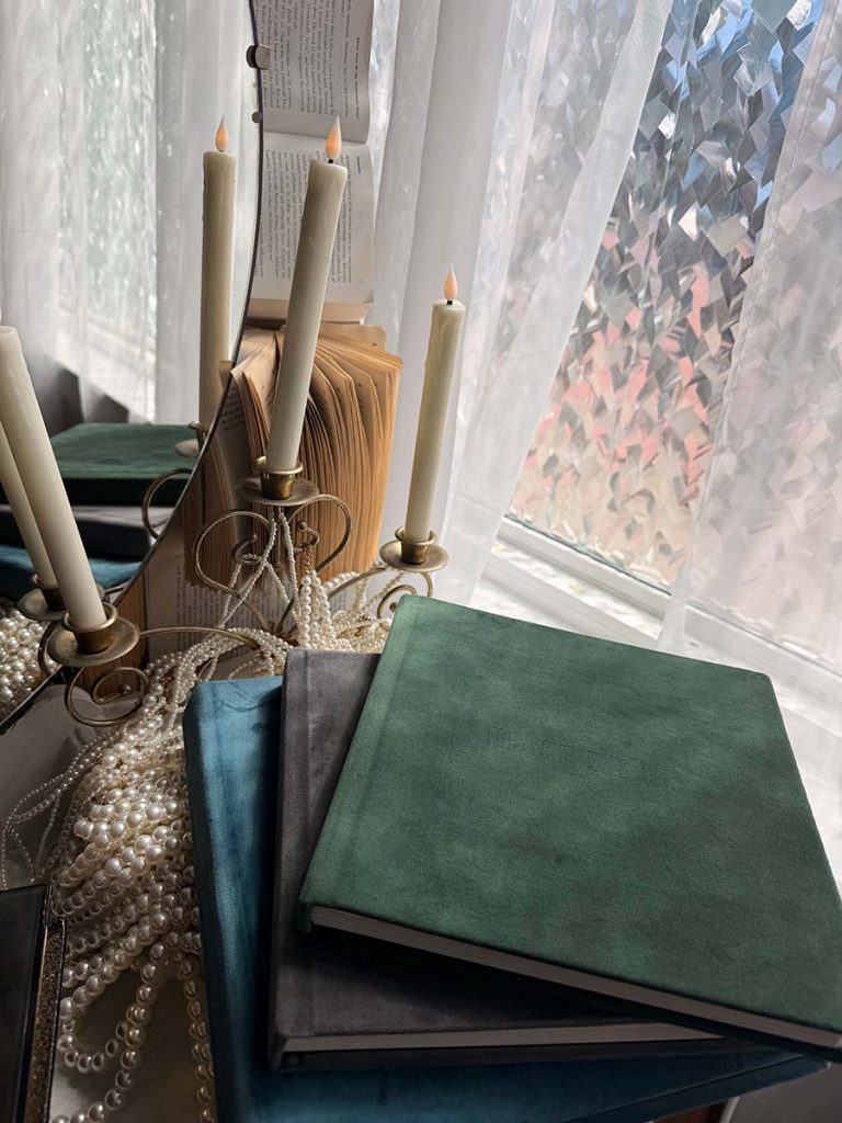 Stack of velvet-covered books beside a mirror reflecting lit candles and an open book. Nearby, a string of pearls adds elegance, with sunlight filtering through a textured window.