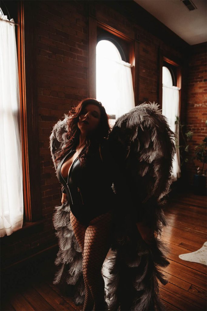 A woman poses in a dimly lit room with large, feathered wings. She wears a black outfit and fishnet stockings, evoking a mysterious, empowering vibe.