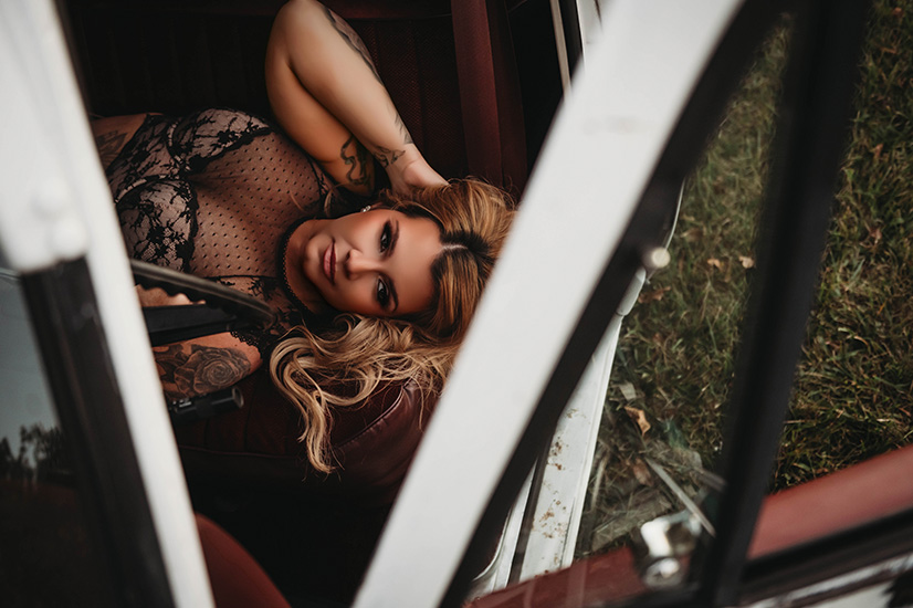 Woman in lace outfit lounges inside a vintage car, gazing upwards with a relaxed expression. The scene conveys a mood of casual elegance.