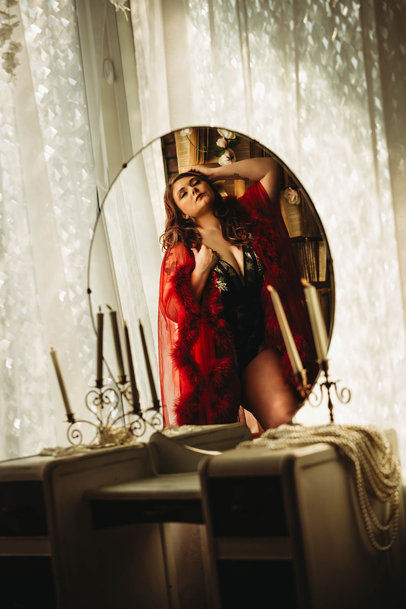 Woman wearing red feathered robe looks upward through decorative oval mirror surrounded by gauzy white curtains, creating a sensual and elegant bedroom portrait.