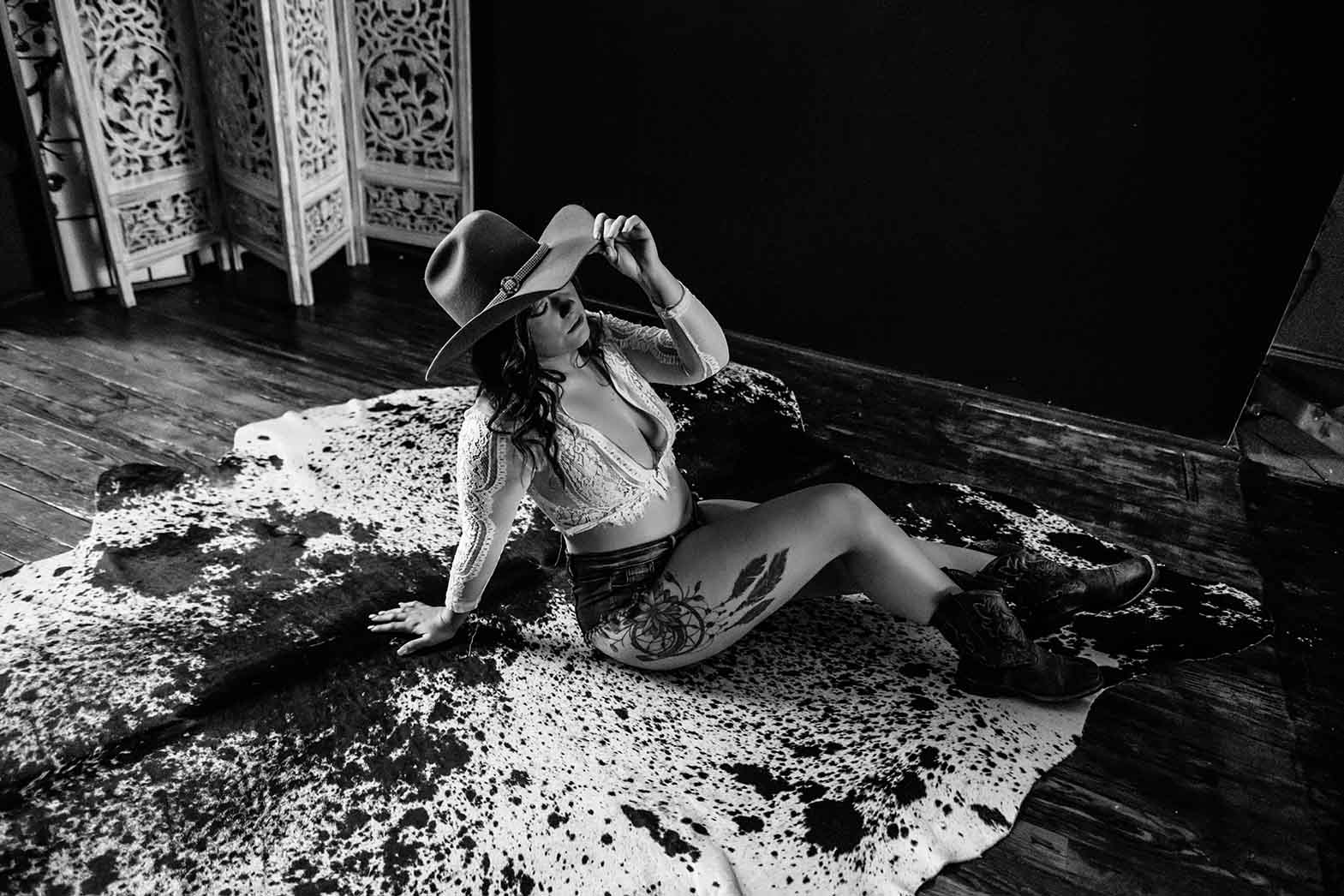 Tattooed woman wearing cowboy hat and lace crop top poses on decorative speckled flooring near carved folding screen, exuding edgy style and artistic presence.