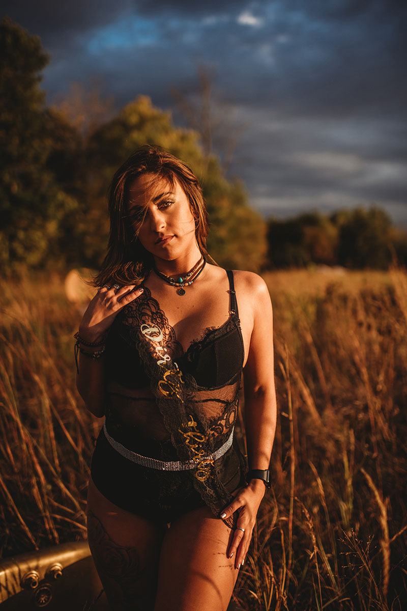 Woman in black lace corset and ornamental choker poses in harvest field during golden hour with dramatic cloudy sky, evoking edgy fashion portrait style.