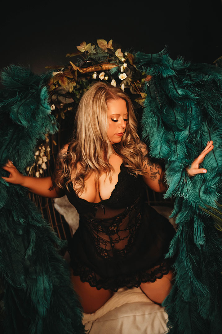 Woman in black lace dress with long highlighted hair stands between emerald feathered wings, gazing downward with warm dramatic lighting and moody atmosphere.