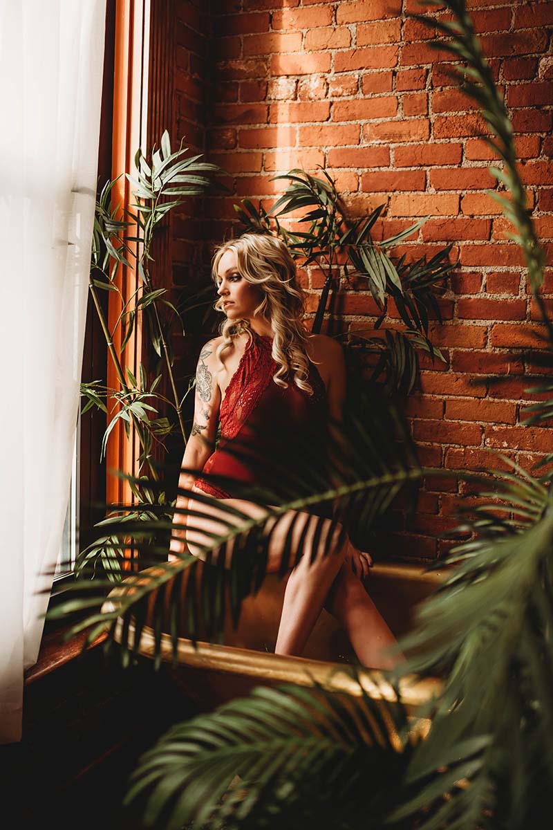 Professional portrait of woman in red dress positioned near exposed brick wall, surrounded by indoor plants with natural window light creating dramatic shadow patterns.